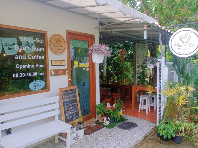 ทางเข้าร้านกับป้าย Khao-Soi Baan Suan and Coffee ม้านั่งสีขาว และกระดานเมนู
