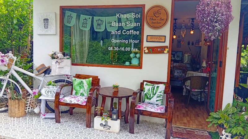 หน้าบ้านสวนของร้าน มีเก้าอี้ไม้ จักรยาน กระดานเมนู และป้าย Khao-Soi Baan Suan ที่หน้าต่าง